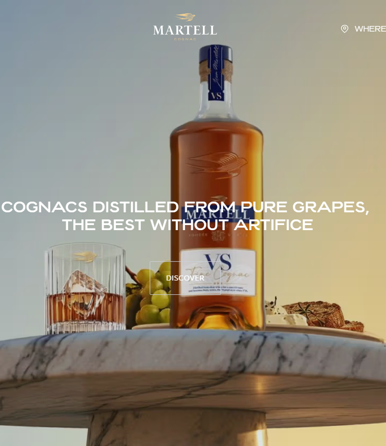 Martell VS Cognac bottle on a marble table with a glass of Cognac on ice, grapes, and cheese in the background, with soft warm lighting highlighting the amber color of the spirit.